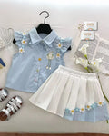 Light blue blouse with floral embroidery and white pleated skirt on a white surface.